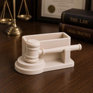 May include: A light-colored wooden gavel and pen holder set. The gavel is resting on a tiered base, with a rectangular pen holder behind it. The set is on a wooden surface, with law books and a scale of justice in the background.