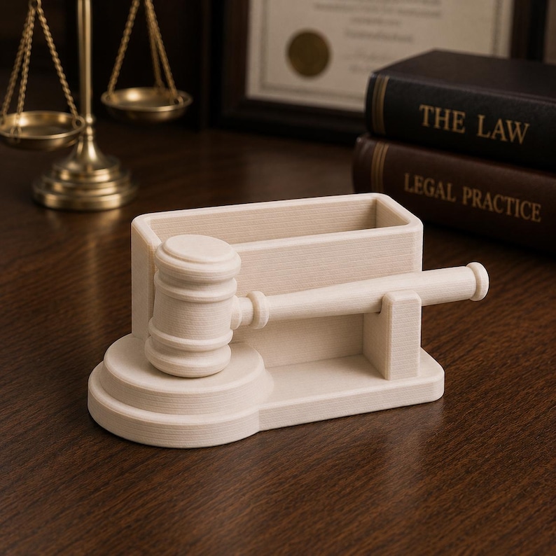 May include: A light-colored wooden gavel and pen holder set. The gavel is resting on a tiered base, with a rectangular pen holder behind it. The set is on a wooden surface, with law books and a scale of justice in the background.