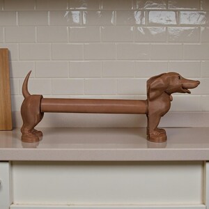 May include: A brown dachshund-shaped paper towel holder. The dog is standing on all fours with its tail up and head facing right. The paper towel roll is held in the dog's body. The holder is made of a solid material and sits on a white countertop.