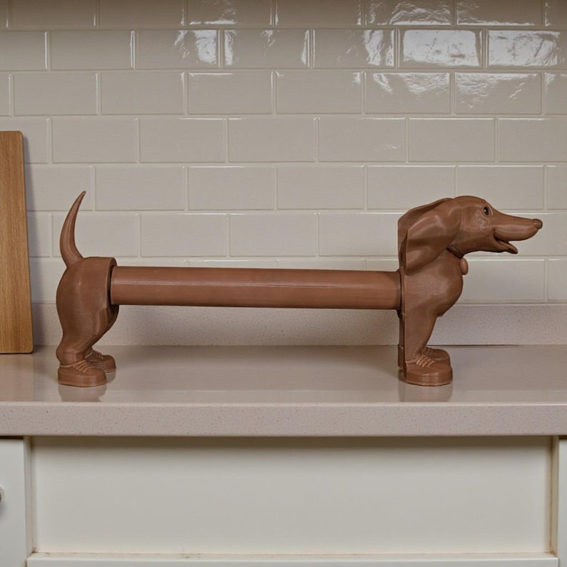 May include: A brown dachshund-shaped paper towel holder. The dog is standing on all fours with its tail up and head facing right. The paper towel roll is held in the dog's body. The holder is made of a solid material and sits on a white countertop.