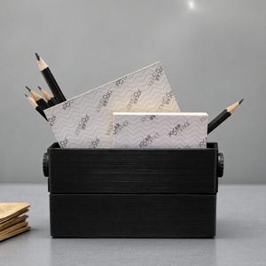 May include: A black wooden desk organizer filled with pencils, notepads, and a small box. The notepads and box have the text "JOCAR OFFICE" printed on them. The pencils are sharpened and ready for use. The organizer is on a gray surface.