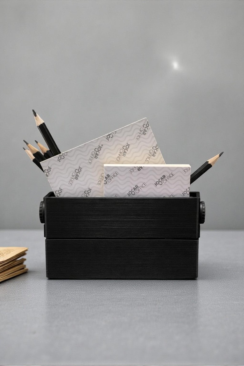 May include: A black wooden desk organizer filled with pencils, notepads, and a small box. The notepads and box have the text "JOCAR OFFICE" printed on them. The pencils are sharpened and ready for use. The organizer is on a gray surface.