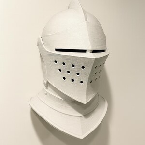 May include: A white knight's helmet with a visor and neck guard. The helmet has a textured surface and a series of small holes on the visor. The helmet is a decorative item, possibly for display or cosplay.