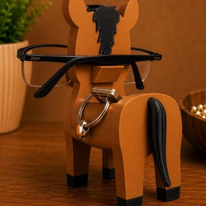 May include: A brown horse-shaped eyeglasses holder with black accents. The holder has a black mane and tail, and is holding a pair of black-framed glasses. A set of keys hangs from a ring on the horse's back.