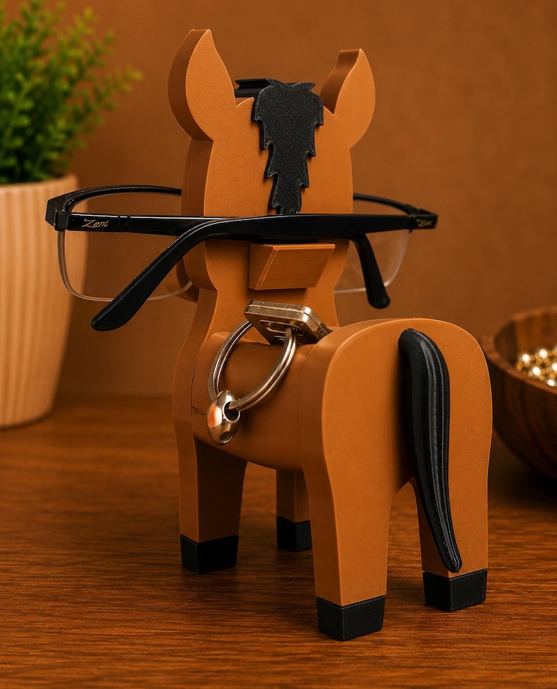 May include: A brown horse-shaped eyeglasses holder with black accents. The holder has a black mane and tail, and is holding a pair of black-framed glasses. A set of keys hangs from a ring on the horse's back.