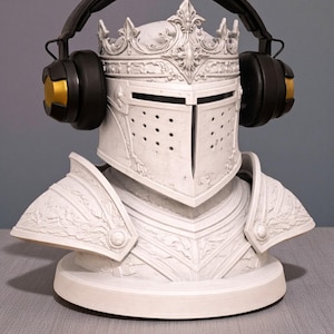 May include: A white bust of a knight's helmet and armor, adorned with a crown and featuring black and gold headphones. The sculpture is detailed with intricate carvings and sits on a round base, creating a unique decorative piece.