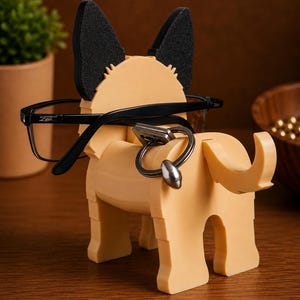 May include: A tan-colored dog-shaped eyeglasses and key holder with black ears. Black-framed eyeglasses rest on the dog's head, and a key ring hangs from the dog's body. The holder is made of a solid material.