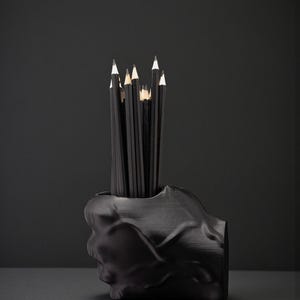 May include: A black skull-shaped pencil holder filled with black pencils. The pencils have white tips and are arranged upright. The holder is made of a matte black material and sits on a dark surface.