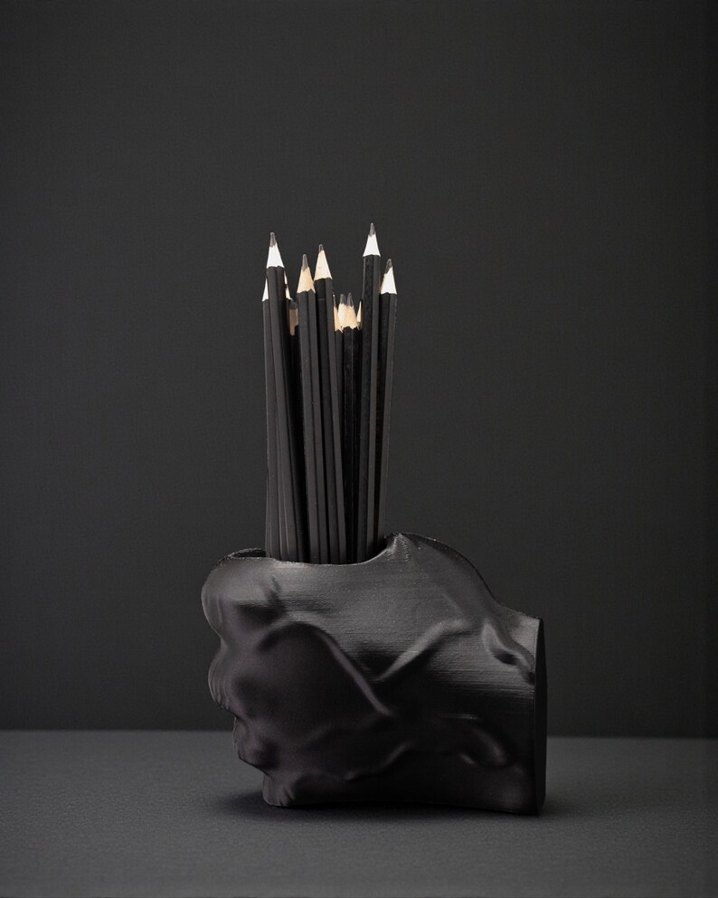May include: A black skull-shaped pencil holder filled with black pencils. The pencils have white tips and are arranged upright. The holder is made of a matte black material and sits on a dark surface.