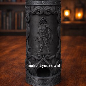 May include: A black cylindrical dice tower with an embossed design featuring a hooded figure. The tower has ornate scrollwork and the text "make it your own!" is visible. The item is on a wooden surface.