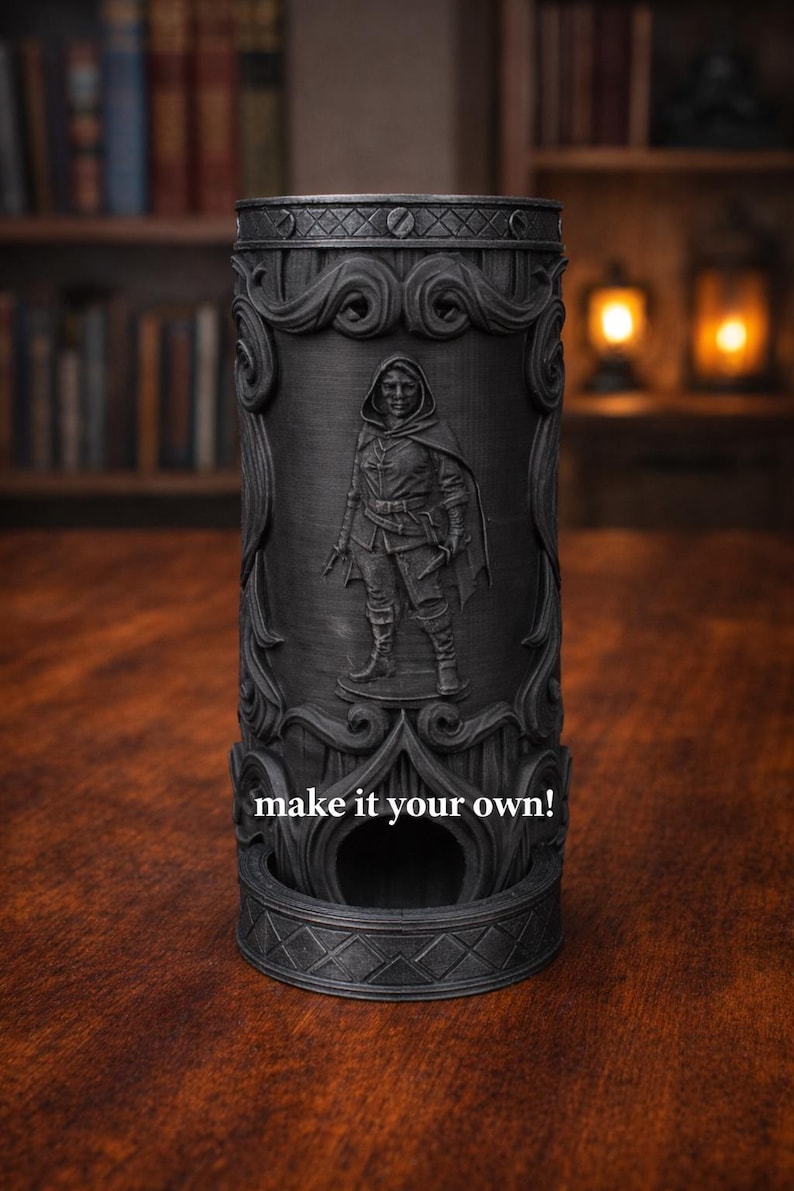 May include: A black cylindrical dice tower with an embossed design featuring a hooded figure. The tower has ornate scrollwork and the text "make it your own!" is visible. The item is on a wooden surface.