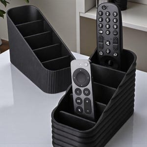 May include: Two black remote control organizers with multiple compartments. One organizer is angled with four sections, and the other has three sections. Two remote controls are placed inside the organizers.