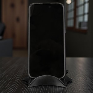 May include: A black smartphone rests on a black, stylized stand. The phone's screen is dark, and the stand has a curved design. The setup is on a dark wooden surface, suggesting a modern aesthetic.