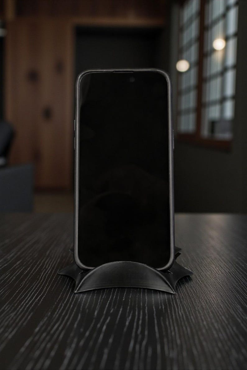May include: A black smartphone rests on a black, stylized stand. The phone's screen is dark, and the stand has a curved design. The setup is on a dark wooden surface, suggesting a modern aesthetic.