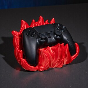 May include: A black video game controller rests in a vibrant red flame-shaped holder. The controller has multiple buttons and two joysticks. The holder is a decorative accessory for gaming enthusiasts.