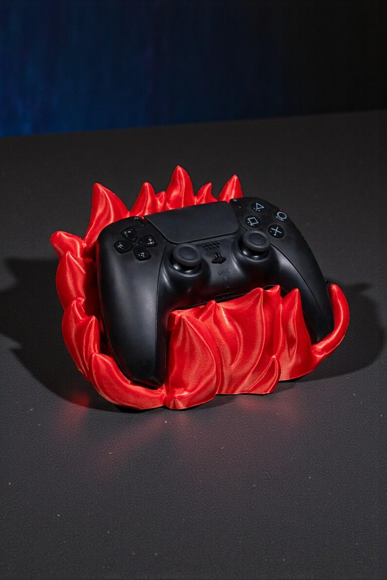 May include: A black video game controller rests in a vibrant red flame-shaped holder. The controller has multiple buttons and two joysticks. The holder is a decorative accessory for gaming enthusiasts.