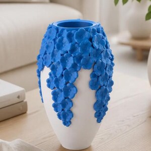 May include: A white vase with a blue floral design. The vase has a rounded shape with a wide opening and is decorated with cascading blue flowers. The vase is sitting on a wooden table.