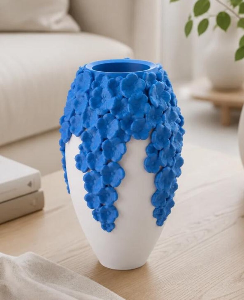 May include: A white vase with a blue floral design. The vase has a rounded shape with a wide opening and is decorated with cascading blue flowers. The vase is sitting on a wooden table.