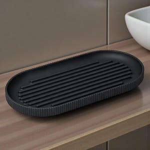 May include: A black, oval-shaped soap dish with a ribbed interior and a textured, ridged border. The dish is made of a matte material and sits on a wooden surface. The design is minimalist and modern.