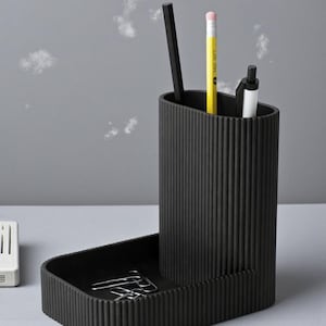 May include: A black desk organizer with a ribbed texture. It features a tall, oval-shaped pen holder and a rectangular tray for small items. Inside the pen holder are a black pencil, a yellow pencil, and a white pen. Paper clips are in the tray.