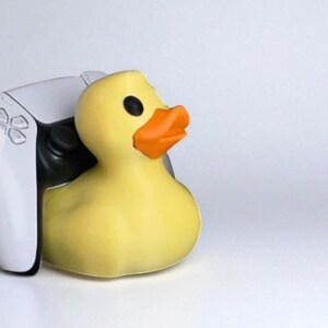 May include: A yellow rubber duck with an orange beak and black eyes rests next to a white and black game controller. The duck is the primary focus, set against a plain white background.
