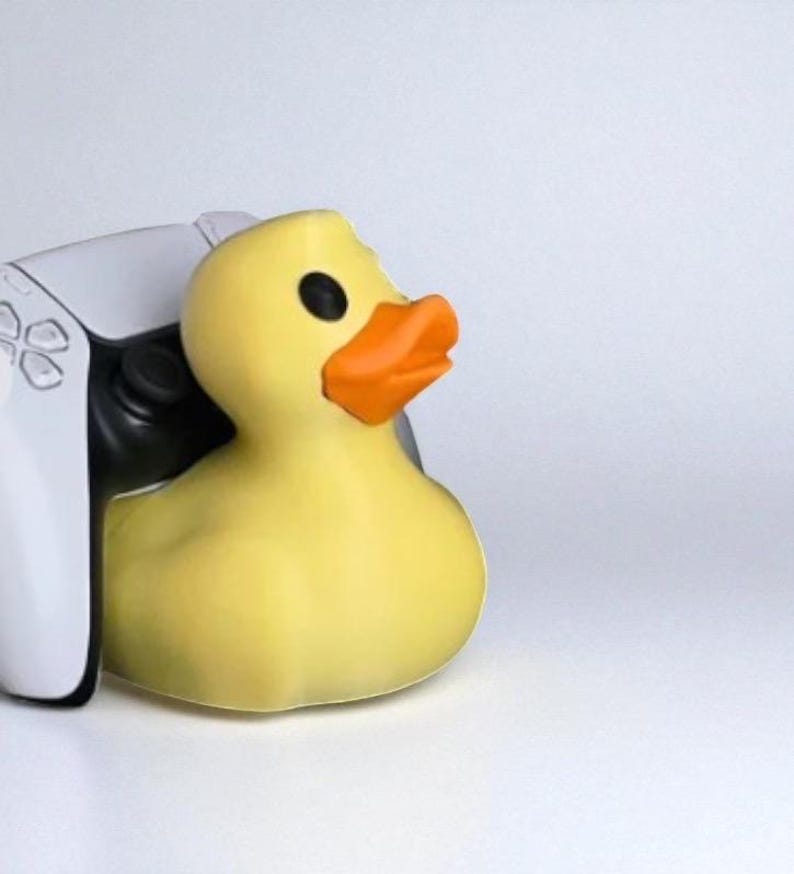 May include: A yellow rubber duck with an orange beak and black eyes rests next to a white and black game controller. The duck is the primary focus, set against a plain white background.