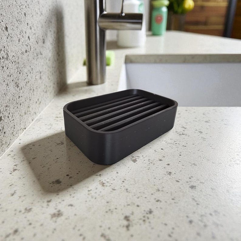 May include: A rectangular black soap dish with a slatted design. The dish is made of a matte material and sits on a speckled countertop near a stainless steel faucet. The soap dish is designed to allow water to drain, extending the life of the soap.