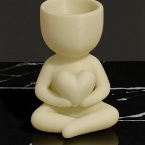 May include: A cream-colored figurine of a person in a seated yoga pose, holding a heart-shaped object. The figurine has a hollowed-out head, possibly for a small plant or decoration. The figurine is made of a smooth material.