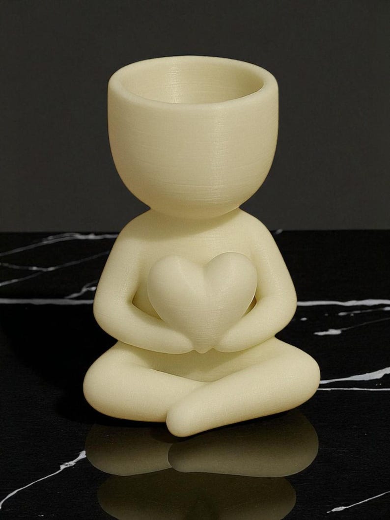 May include: A cream-colored figurine of a person in a seated yoga pose, holding a heart-shaped object. The figurine has a hollowed-out head, possibly for a small plant or decoration. The figurine is made of a smooth material.