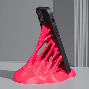 May include: A vibrant pink phone stand with a unique, abstract design. The stand supports a black smartphone, creating a striking contrast. The stand's form appears to be flowing or melting, adding a modern touch to the desk accessory.