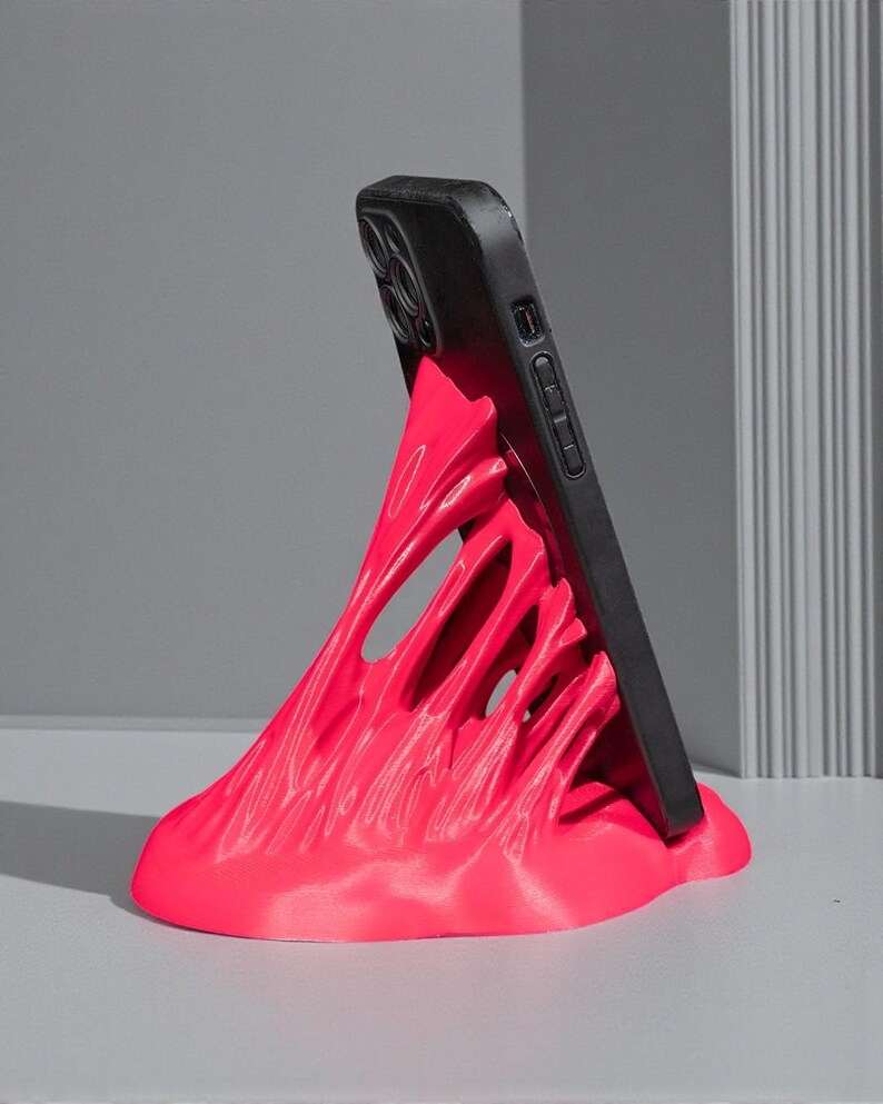 May include: A vibrant pink phone stand with a unique, abstract design. The stand supports a black smartphone, creating a striking contrast. The stand's form appears to be flowing or melting, adding a modern touch to the desk accessory.