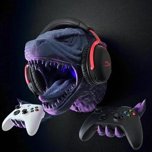 May include: A blue and purple dinosaur head-shaped headset holder with black and red headphones. Two game controllers, one white and one black, are held in the dinosaur's hands. The background is black.