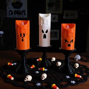 May include: Three Halloween-themed LED candles in orange and white, each with a carved face design. The candles sit on black candle holders, surrounded by candy corn, small skulls, and decorative accents on a black lace doily.