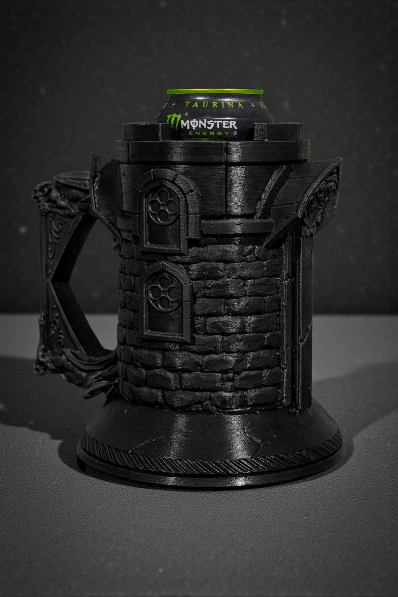 May include: A black, 3D-printed mug shaped like a castle tower, with a handle and decorative details. A green and black Monster Energy drink can sits inside the tower. The mug has a textured brick pattern.