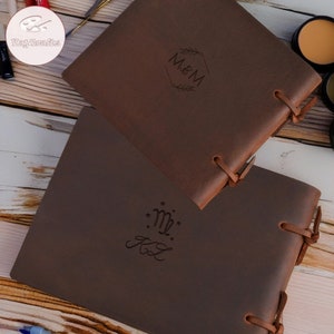 Może przedstawiać: Dwa brązowe sk&oacute;rzane dzienniki z personalizowanymi grawerami. G&oacute;rny dziennik ma inicjały "M&M" w sześciokątnym ramie z liśćmi. Dolny dziennik ma znak zodiaku Panna z inicjałami "KL" pisanymi kursywą.