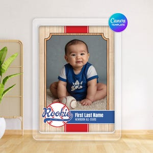 Puede incluir: Foto de bebé en un marco acrílico transparente. La foto presenta a un bebé con un body azul y blanco, una pelota de béisbol y el texto "Rookie of the Year" y "Newborn All-Stars". El marco tiene un fondo de vetas de madera con detalles en rojo.