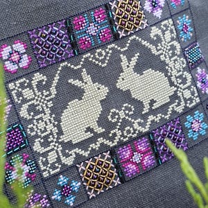 May include: A cross-stitch embroidery design featuring two white rabbits in a gray frame with a floral border. The border is made up of small squares with different colored floral patterns and beads.