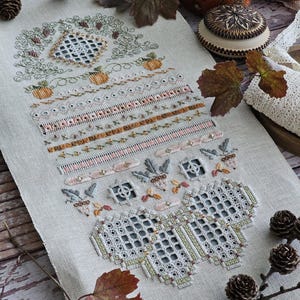 以下が含まれることがあります： 秋をテーマにした、詳細なクロスステッチのサンプル。デザインには、カボチャ、花の模様、幾何学的な形が含まれており、すべてオレンジ、緑、ベージュの色合いで表現されています。サンプルはリネンの背景に表示され、装飾的な要素があります。