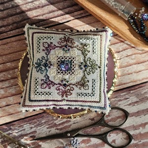 May include: A square, embroidered pincushion with a floral design in shades of purple, green, and blue. The pincushion is surrounded by a beaded border. A pair of vintage scissors and a small tray with beads are also visible.
