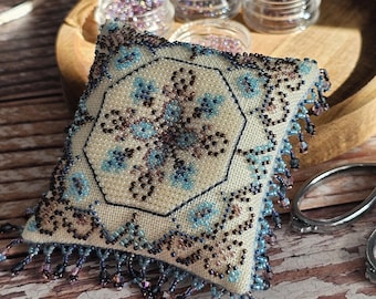 May include: A small, square, beige pincushion with a blue and brown beaded design. The pincushion has a fringe border and is sitting on a wooden surface.