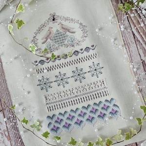 Op de afbeelding: Een witte linnen stof met een delicaat borduurwerk met vogels, harten, sneeuwvlokken en geometrische patronen in tinten paars, blauw en grijs.