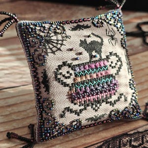 Könnte beinhalten: Ein quadratisches, kreuzgesticktes Halloween-Kissen mit einem schwarzen Kater, Spinnweben und einem Kessel-Design. Die Kanten sind mit Perlen verziert. Das Kissen ist etwa 7,6 cm groß.