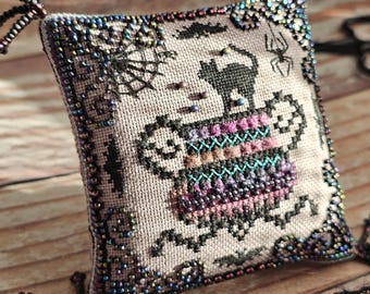 May include: A square, cross-stitched Halloween-themed pillow with a black cat, spiderwebs, and a cauldron design. The edges are embellished with beads. The pillow is approximately 3 inches square.