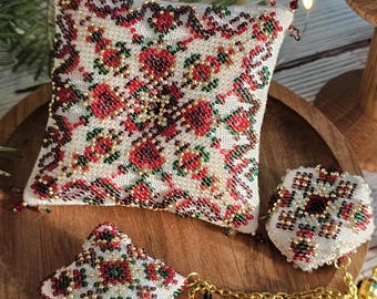 May include: A collection of beaded ornaments with a floral pattern in red, green, and gold on a white background. The ornaments include a small pillow, a pendant, and a round charm, all displayed on a wooden tray.