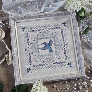 May include: A square framed cross-stitch embroidery featuring a central hummingbird surrounded by intricate geometric borders, floral motifs, and small crystal embellishments, with four smaller hummingbirds in the corners of the design.
