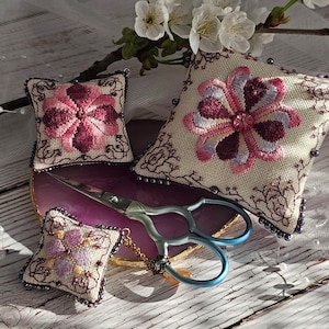 May include: Three small embroidered cushion covers with floral designs in shades of pink and purple. The cushions are surrounded by white pearl beads and a pair of blue scissors on a purple agate coaster.