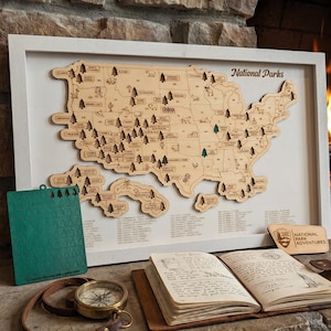 May include: A framed wooden map of the United States featuring National Parks, a green rectangular card with tree patterns, a brass compass with a brown leather strap, and an open journal with handwritten notes and a National Park Adventures badge.