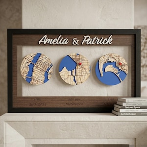 May include: A framed artwork with three circular maps, each with a blue background and a red pin. The frame includes the names "Amelia & Patrick" and dates below the maps. The artwork is displayed on a white surface.