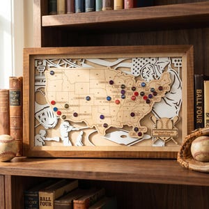 May include: A framed wooden map of the United States featuring baseball-themed cutouts of players, a glove, and a stadium. The map includes small circular markers placed on various states and a plaque labeled Michael Family Stadium Map.