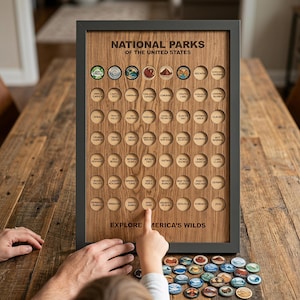 May include: A rectangular wooden board in a dark grey frame titled NATIONAL PARKS OF THE UNITED STATES with rows of circular cutouts for tokens. The board features the text EXPLORE AMERICA'S WILDS and includes many colourful circular park tokens.